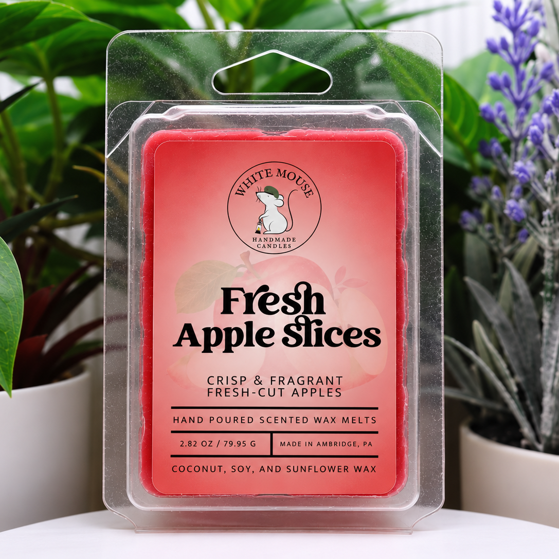 A packaged wax melt labeled 'Fresh Apple Slices' by White Mouse Candles, shown in a clear blister pack against a background of potted green and purple plants.
